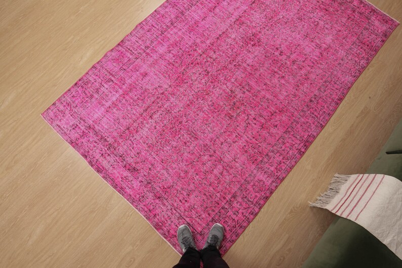 May include: A rectangular, hot pink rug with a distressed, vintage appearance. The rug has a darker border and is placed on a light wood floor. The rug's dimensions are not visible.