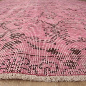 May include: A large area rug featuring a vibrant pink base color with intricate, dark brown floral and geometric patterns woven throughout. The rug has a distinct border design and a short, light-colored fringe along the bottom edge.