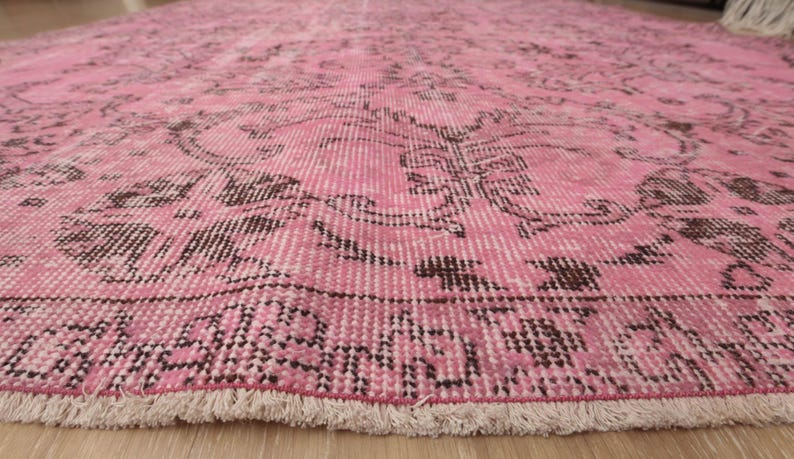 May include: A large area rug featuring a vibrant pink base color with intricate, dark brown floral and geometric patterns woven throughout. The rug has a distinct border design and a short, light-colored fringe along the bottom edge.
