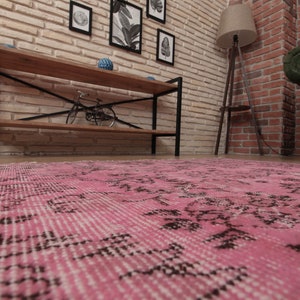 May include: A rectangular area rug featuring a vibrant pink base color with intricate, faded black floral and geometric patterns distributed across the surface. The rug has a low-pile texture and visible fringe detailing along the short edges.