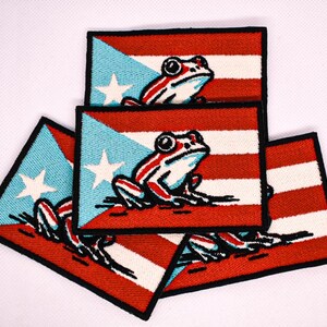 May include: Rectangular embroidered patches featuring the Puerto Rican flag design with a frog illustration. The patches have a red and white striped background, a blue triangle with a white star, and a frog with red and white stripes.