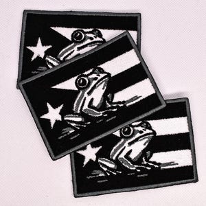 May include: Three embroidered patches featuring a black and white design. Each patch displays a flag with a white star and a frog illustration. The patches are rectangular with a black border.