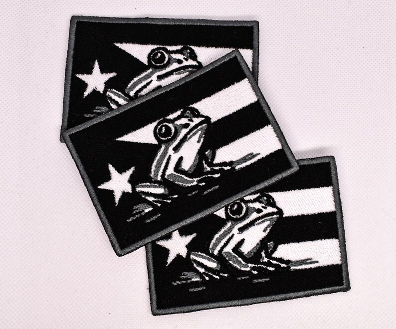 May include: Three embroidered patches featuring a black and white design. Each patch displays a flag with a white star and a frog illustration. The patches are rectangular with a black border.