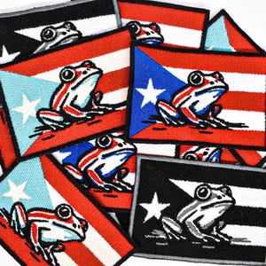 May include: A collection of embroidered patches featuring the Puerto Rican flag and a frog design. The patches display the flag's colors: red, white, and blue, with a white star and a frog in the center. The frog is white with red and blue accents.