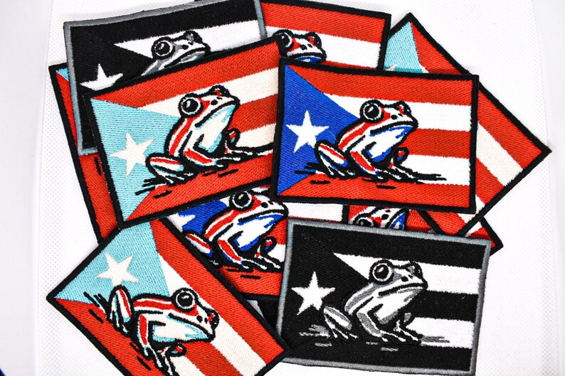 May include: A collection of embroidered patches featuring the Puerto Rican flag and a frog design. The patches display the flag's colors: red, white, and blue, with a white star and a frog in the center. The frog is white with red and blue accents.