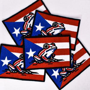 May include: A collection of embroidered patches featuring the Puerto Rican flag design. Each patch has a blue triangle with a white star, red and white stripes, and a frog illustration. The patches have a black border.