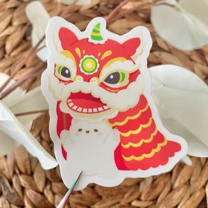 May include: A white sticker with a red and gold lion dance costume. The lion has a green and yellow face with a red mouth and white teeth. The lion is holding a white cat with pink cheeks.
