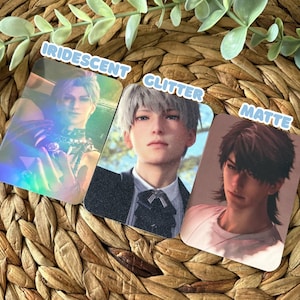 May include: Three rectangular cards featuring anime-style character portraits. The left card has an iridescent finish showing a character with light hair. The middle card has a glitter finish showing a character in a suit. The right card has a matte finish showing a character with dark hair.