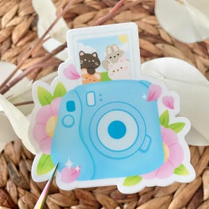 May include: A blue and white instant camera with a photo of two cartoon animals inside. The camera is surrounded by pink flowers and green leaves.