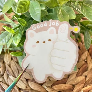 May include: White sticker with a cartoon cat giving a thumbs up. The cat has brown eyes and a pink nose. The sticker says "Good Job" in pink letters.