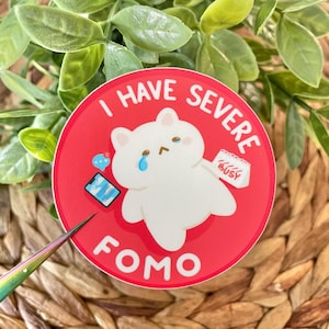 May include: A red and white round sticker with a white cartoon cat with a tear on its cheek. The sticker says "I have severe FOMO" with a calendar that says "BUSY" in the background.