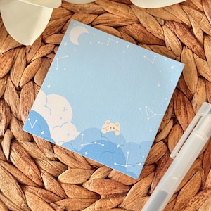 May include: A square, pale blue sticky note with a celestial design. The design features white clouds, constellations, a crescent moon, and a small, light brown bear peeking over the clouds. A white pen rests on the right side.