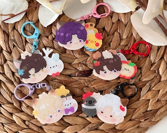 May include: Six colorful cartoon character keychains with different animal and character designs. Each keychain features a small character with a larger character behind it. The keychains are on a woven brown background.