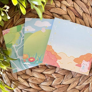 Op de afbeelding: Drie pastelkleurige plakbriefjes met verschillende ontwerpen. De eerste plakbriefje heeft een groene heuvel met een witte kat, de tweede plakbriefje heeft een blauwe lucht met witte sterren en de derde plakbriefje heeft een roze en oranje lucht met een witte kat.
