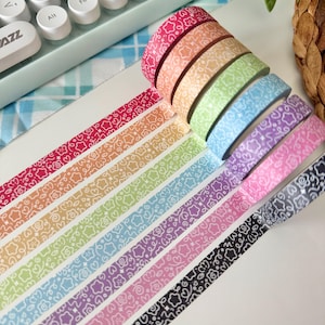 May include: A selection of rainbow-coloured washi tapes, each featuring a white floral and swirl design. The tapes are stacked and unrolled, displaying a range of colours and patterns. Ideal for crafting and adding a decorative touch to projects. Each roll is approximately 1.5 cm wide.