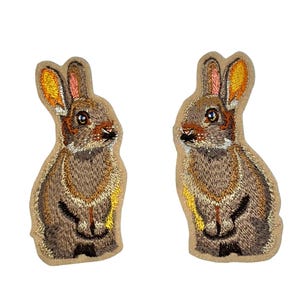 May include: Two embroidered rabbit patches. The rabbits are brown with tan and yellow detailing. The patches are set against a tan backdrop. The patches feature detailed embroidery.
