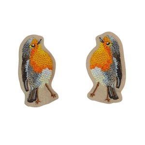 May include: Two embroidered patches of a European robin. The birds have orange breasts, grey and brown wings, and are set against a tan background. The patches are detailed with various thread colours.