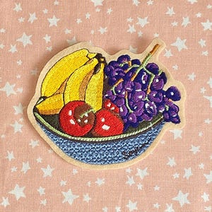 May include: Embroidered patch featuring a bowl of fruit. The bowl contains yellow bananas, purple grapes, and red cherries. The bowl is blue with a textured design, and the patch has a beige border.