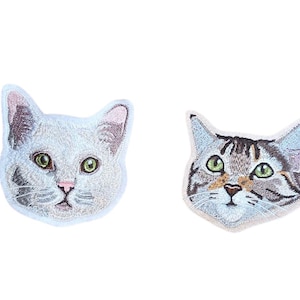 Puede incluir: Dos parches bordados con caras de gato. Un parche presenta un gato blanco con ojos verdes y detalles rosas. El otro parche representa un gato atigrado con ojos verdes y marcas marrones y grises. Ambos parches tienen un borde blanco.