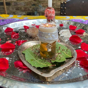 Op de afbeelding: Een zilveren dienblad met een witte kaars, een rode kaars en een glazen pot met een witte kaars erin. Het dienblad is omringd door rozenblaadjes en laurierbladeren.