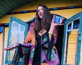 May include: A woman wearing a long, black and colorful patterned coat with fringe, black tights, and silver shoes stands in front of a yellow and green wooden structure. She is holding a small blue and white box in her right hand.