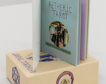 Puede incluir: Un libro cerrado titulado "Aetheric Tarot" con una cubierta azul claro y una foto de una mujer en un círculo en la portada. El libro está sentado encima de una caja con un diseño de calavera y ojo en la parte delantera. La caja está etiquetada como "Aetheric Tarot" en letras azules.