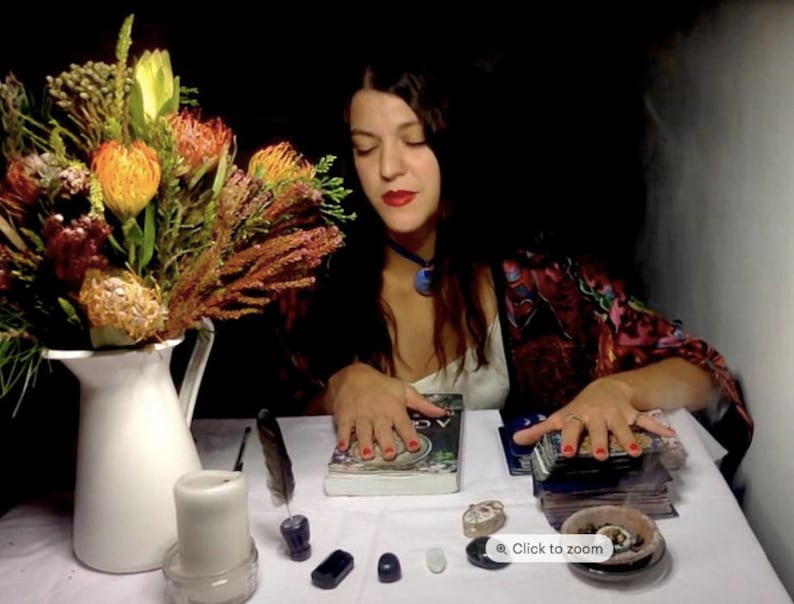 May include: A woman with long dark hair sits at a table with a white tablecloth. She is wearing a red and black patterned robe and has red nail polish. In front of her are a book, a deck of tarot cards, a white candle, and a small bowl with incense.
