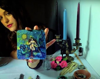 May include: A woman holds a tarot card with a blue, green, and yellow illustration of a woman standing on a fish. The card is surrounded by candles, flowers, and other objects on a white table.