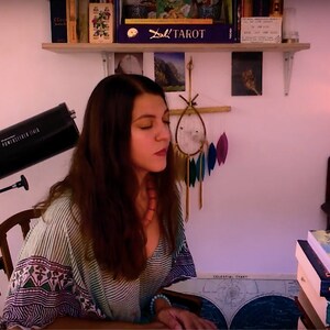 Puede incluir: Una mujer con cabello largo y castaño se sienta en una mesa con un telescopio en el fondo. Lleva una camisa a rayas y un collar. Hay libros sobre la mesa, incluyendo "The Black Sun", "Dispelling Wetiko" y "The Great Cosmic Mother". También hay velas sobre la mesa.