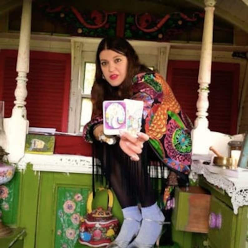 May include: A woman sits in a colorful, decorated wagon and holds a tarot card with a white unicorn design. The card is surrounded by a white border and has a pink background.