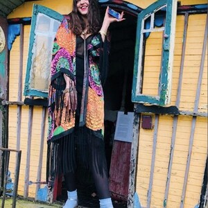 May include: A woman wearing a long, black and colorful patterned kimono with black fringe. She is standing in front of a yellow wooden building with a green door. She is holding a small blue object in her right hand.