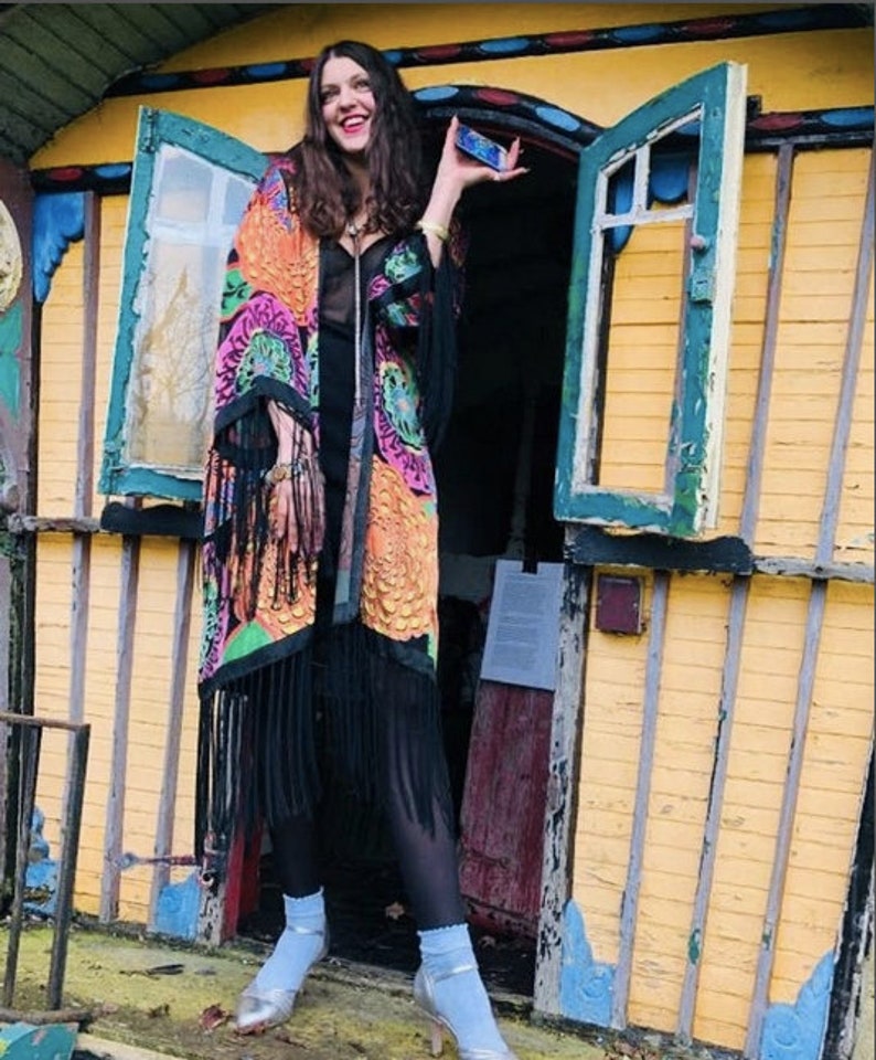 May include: A woman wearing a long, black and colorful patterned kimono with black fringe. She is standing in front of a yellow wooden building with a green door. She is holding a small blue object in her right hand.