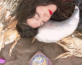 May include: A woman sleeps with her head resting on a white crescent moon-shaped pillow. The pillow has a black eye drawn on it. The woman is wearing a black shirt and has long dark hair. A gold foil blanket is visible in the background.