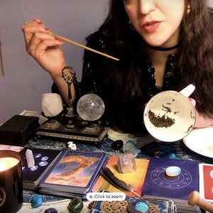 May include: A woman is holding a white teacup with tea leaves in it. The teacup is on a white saucer. The woman is sitting at a table with a variety of crystals, candles, and tarot cards. The table is covered in a dark blue tablecloth. The woman is wearing a black and white floral dress. The image is a close-up of the woman's hands and the teacup.