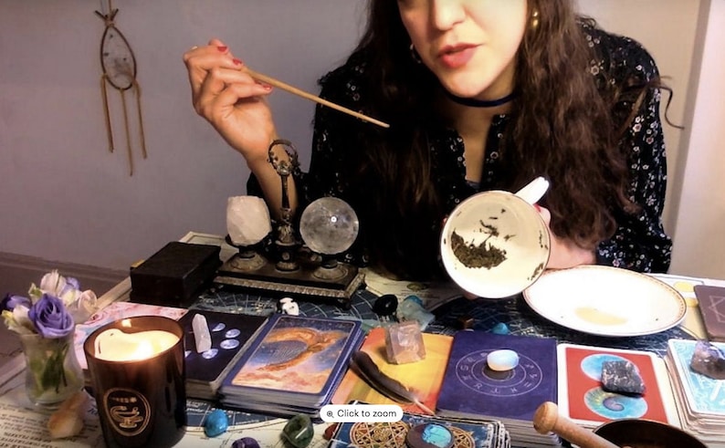 May include: A woman is holding a white teacup with tea leaves in it. The teacup is on a white saucer. The woman is sitting at a table with a variety of crystals, candles, and tarot cards. The table is covered in a dark blue tablecloth. The woman is wearing a black and white floral dress. The image is a close-up of the woman's hands and the teacup.