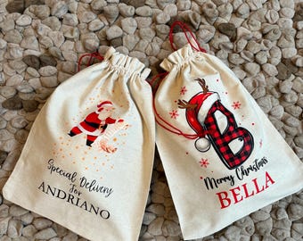 Puede incluir: Dos bolsas de regalo de lona blanca con cordones de extracción rojos. Una bolsa tiene una ilustración de Papá Noel y el texto "Special Delivery for ANDRIANO". La otra bolsa tiene una letra "B" de cuadros rojos y negros con un reno y el texto "Merry Christmas BELLA".