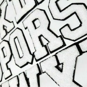 May include: Close-up of white varsity letter patches with black borders. The letters are in a bold, block style, and the patches have a textured, fuzzy appearance. The letters are arranged at various angles.