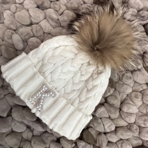 May include: White knitted beanie with a faux fur pom pom and a sparkly letter K on the front.