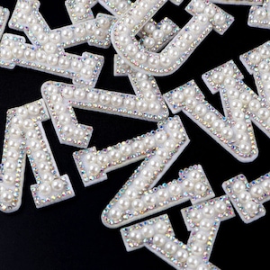 May include: A collection of white pearl-embellished letters, including the letters A, M, W, and Y. The letters are made of a white fabric base and are decorated with white pearls.