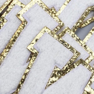 Könnte beinhalten: Mehrere weiße Filzbuchstaben mit goldfarbenem Glitzerbesatz. Die Buchstaben sind übereinander gestapelt und ergeben das Wort "RAW".