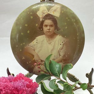 May include: A vintage photo of a young girl with a white bow in her hair, framed in a round, gold-toned frame. The photo is set on a decorative stand with a pink flower.