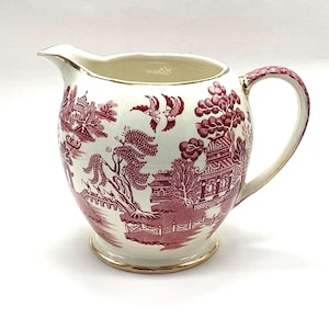 May include: A cream-colored ceramic creamer pitcher featuring a deep red transferware pattern of pagodas, willow trees, and birds. The pitcher has a gold-trimmed rim, a gold-trimmed base, and a handle with a matching red patterned design.