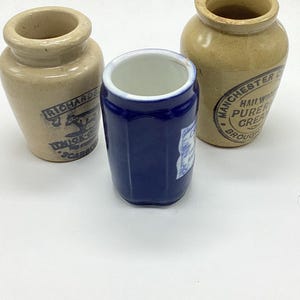 May include: Three antique stoneware jars. Two are tan with blue text and graphics, and one is a deep blue with a white interior. The jars have a cylindrical shape with a wide opening at the top. The jars are likely from the early 20th century.