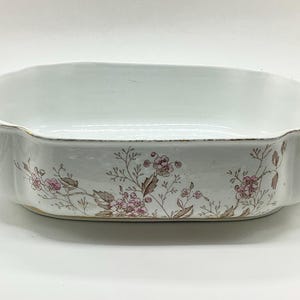 May include: A white ceramic serving dish with a rectangular shape and scalloped edges. The dish features floral patterns in pink and brown, with gold trim. It has two ornate handles on either side, suitable for serving food.