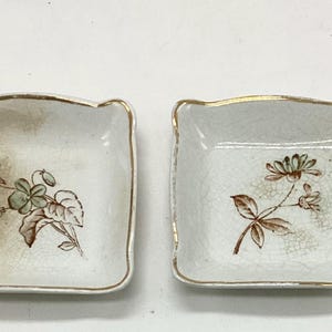 May include: Two square ceramic dishes with rounded corners and gold-painted rims. Each dish features a white base with a visible crackle glaze finish and a central botanical illustration of green and brown flowers with leaves.