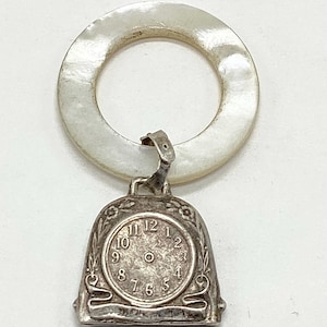 May include: A vintage silver-toned baby rattle with a clock face design. The rattle features a circular, pearlescent handle and a bell-shaped charm with a clock face displaying Roman numerals. The clock charm is adorned with floral accents.