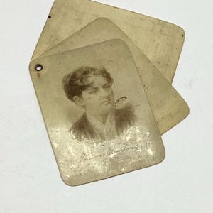 May include: Three rectangular, off-white vintage cards with rounded corners, layered and fanned out. The top card features a sepia-toned portrait of a person with dark hair and the handwritten text "Frances Cleveland" at the bottom, with a metal eyelet.