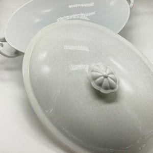 May include: A white, oval-shaped serving dish with a matching lid. The lid features a decorative, flower-shaped handle. The dish has two handles on either side. The surface of the dish and lid have a crackle glaze.