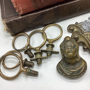 May include: Five antique brass drawer pulls with a ring design and a small screw. The pulls are arranged in a semi-circle on a white surface. There is also a small brass figurine of a person with a crown on their head.