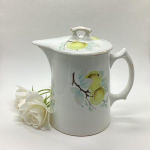 May include: White ceramic pitcher with lid, featuring a handle and spout. The pitcher is decorated with a yellow apple and leaf design. The rim and handle have gold trim. A white rose is next to the pitcher.